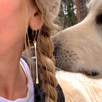 A close-up of a woman wearing Upstream Paddle Earrings, featuring a gold finish, with a curious pet in the background.