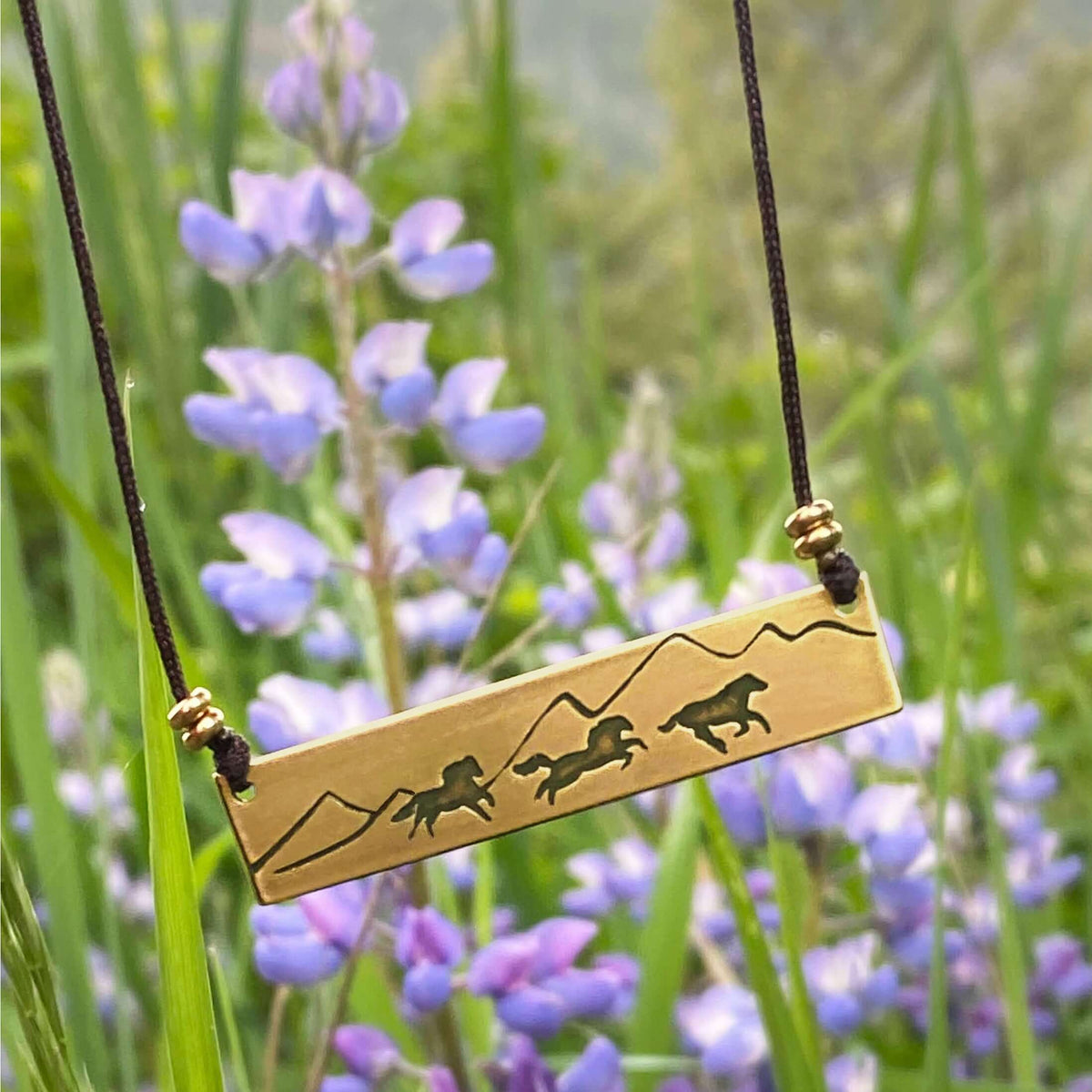 Wild Horses Necklace featuring a brass pendant with etched wild horses and mountains, surrounded by blooming purple flowers.