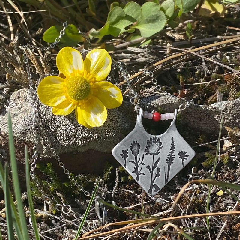 Wildflower Box Necklace with Indian Paintbrush design, silver finish, and pinks stones beside a yellow wildflower.